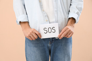 Fototapeta na wymiar Young man holding paper with word SOS on beige background, closeup. Prostate cancer awareness concept