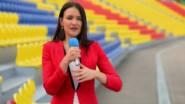 Woman journalist gestures and speaks in microphone near empty seats on sports stadium