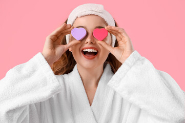 Beautiful young woman in bath headband with heart-shaped sponges on pink background