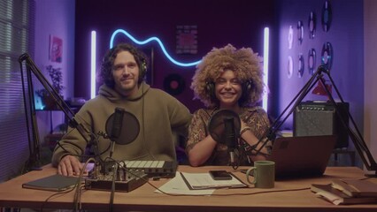 Full medium portrait of smiling diverse male and female hosts of podcast or online show ready for recording at neon lit home studio at night