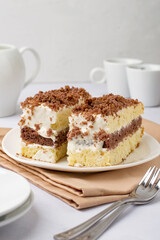Two pieces of cake on a plate, coffee cups, milk jug, cutlery, napkin on a light background.
