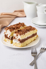 Two pieces of cake on a plate, coffee cups, milk jug, cutlery, napkin on a light background.