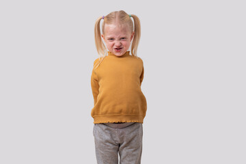 A grimacing little girl in a yellow sweater stands with her hands behind her back against a gray...