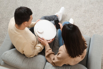 Young couple with modern robot vacuum cleaner and mobile phone sitting on sofa in living room, top...