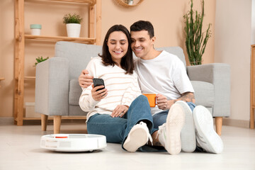 Young couple using mobile phone for remote control of modern robot vacuum cleaner in living room