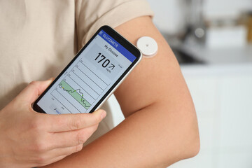 Diabetic woman with glucose sensor using mobile phone for measuring blood sugar level in kitchen,...