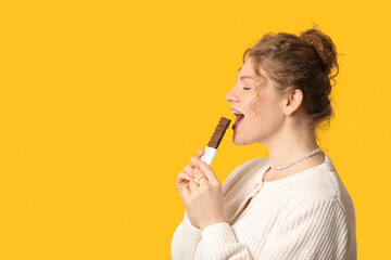 Beautiful young woman eating sweet chocolate bar on yellow background