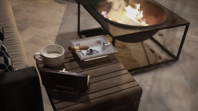 Small wooden cigar table with humidor, ashtray with burning cigar, cup of coffee near sofa and portable fireplace with fire