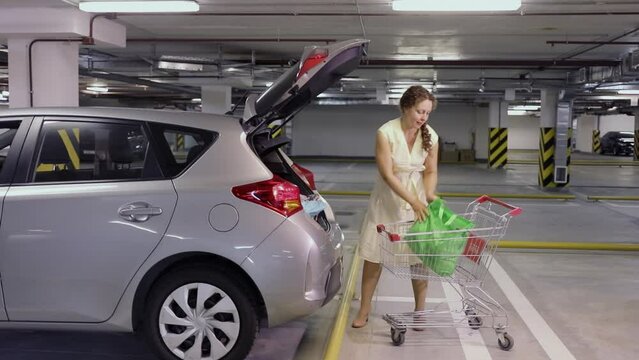 Woman moves bags from shopping cart to car boot at underground parking. Slow motion
