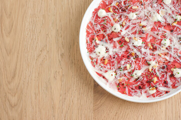 Top view of Beef Carpaccio appetizer with grated Parmesan cheese and pine nuts on white plate with light wood background