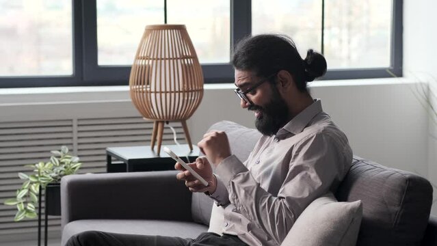 Positive Indian man using phone and laughing relaxing on sofa at home after hard work day. Reading emails, playing video games, leisure time, resting at home.