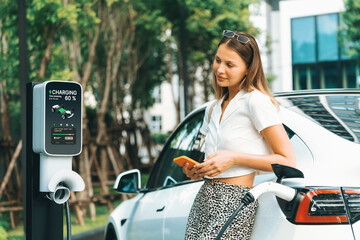 Young woman travel with EV electric car charging in green sustainable city outdoor garden in...
