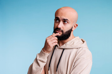 Arab man making decision, touching chin thoughtfully and looking away. Young bald bearded person with pensive facial expression contemplating, thinking about problem solving