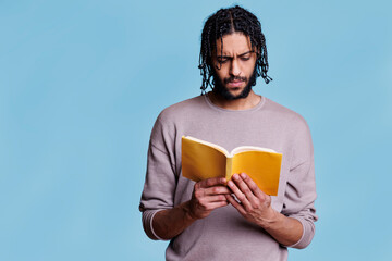 Concentrated arab man reading paperback book with yellow cover. Focused young person with thoughtful expression studying literature, holding softcover textbook on blue background