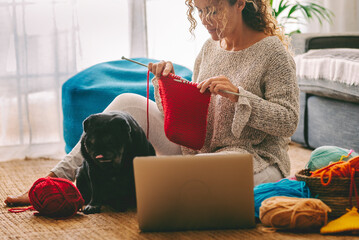 Woman sitting on the floor crocheting a ball of red wool with her black pug dog next to her....