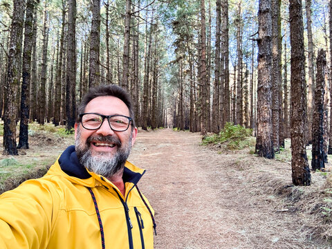 Cheerful adult happy man taking selfie picture in the woods enjoying outdoors leisure activity in national forestal park alone. Adventure and lifestyle people. High trees in background. Sustainability
