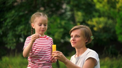 Happy mother with joyful daughter blows bubbles in park among trees on vacation. Preschooler girl waits till mother blows bubbles in meadow at weekend. Mother and child spend time blowing bubbles