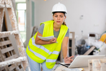 Young woman builder holding documentation, using laptop while working in apartment.