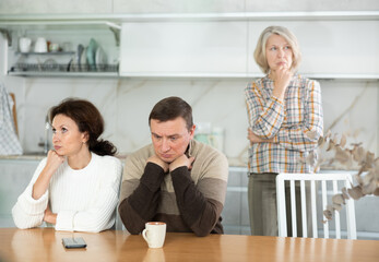 Angry middle-aged man and woman sitting at table and upset old woman standing behind in the kitchen