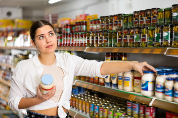 in supermarket, young woman thoughtfully chooses mayonnaise.