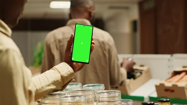 African american storekeeper holding phone with greenscreen in local grocery store, having organic products on sale. Vendor presents isolated chromakey display on smartphone, mockup layout. Camera A.