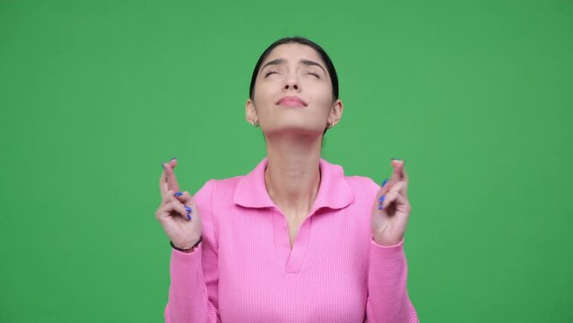 Optimistic young Caucasian woman portrait with crossed fingers in hope, showing desire and strong faith, praying and making wish on green background.