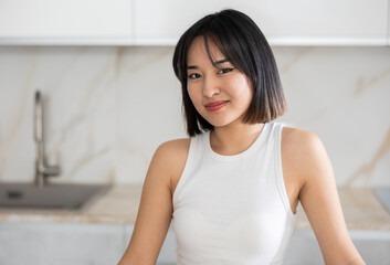 Smiling young Asian girl standing happily at table in the kitchen with white interior