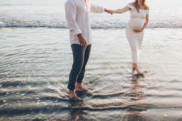 close up crop photo of a pregnant maternity couple holding hands looking at each other standing in the water at sunset on the beach