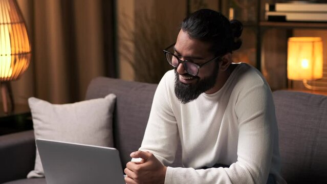 Indian man spends free evening time relaxing on the sofa in the evening living room, communicates via video and using a laptop, laughs, is captivated by online entertainment.