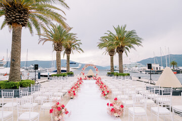 Rows of chairs line a path decorated with red flowers in front of a wedding arch on the pier