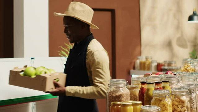 Business owner restocking crates of fresh fruits and vegetables, presenting organic locally grown produce to woman shopping at store. African american people chatting about vegan food. Camera A.
