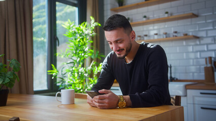 Smiling man sit in kitchen surfing internet on mobile phone, reading media news, scrolling social...