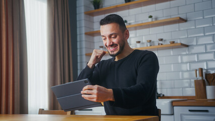 Overjoyed man playing a video game on a tablet computer while sitting in kitchen at home. Excited concentrated man engrossed in interesting racing game, doing winner gesture