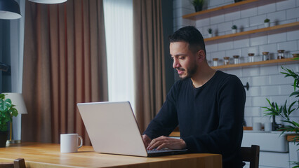 adult man sit in the kitchen typing on laptop computer chatting with friends, enjoying surfing internet, using social media