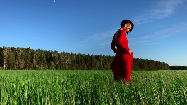 Young woman model in scarlet dress flowing in the wind. Stock clip. Brunette woman with curly hair in fild with forest and blue sky on the background.