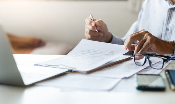 Asian businessman reviewing document reports at office workplace with computer laptop. legal expert, professional lawyer reading and checking financial documents or insurance contract before sign - Powered by Adobe