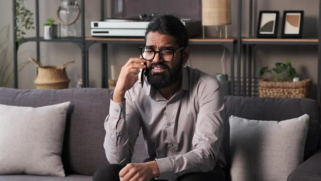 Serious Indian businessman talking on the smartphone at home sitting at sofa. Work at home, remote business consulting, communication, conversation concept.