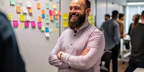 Smiling and laughing manager in a bright spacious office space, with sticky notes on wall. He is a young beard man, plus size, wearing a pink shirt, a good and gentle man. Great company at work.