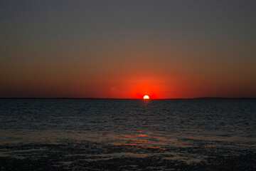 Sunset over the sea against the background of the coast. Evening on the sea calm in clear weather. The disk of the sun in the evening sky over the sea.