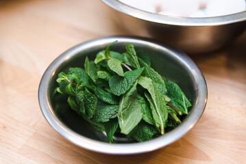 Fresh Mint Leaves in Stainless Steel Bowl. A stainless steel bowl filled with vibrant fresh mint leaves on a wooden kitchen surface, highlighting natural ingredients.