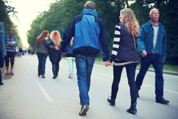 Couple, back and holding hands while walking among trees and crowds along road in New Zealand. Rear...