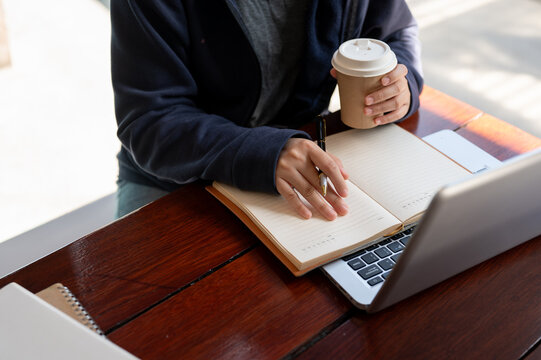 A cropped shot of a woman in a hoodie is sipping coffee while working at a table in a cafe.