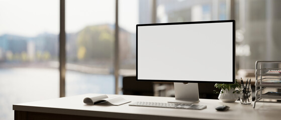 A modern office features a white-screen computer mockup and accessories arranged on a table. - obrazy, fototapety, plakaty