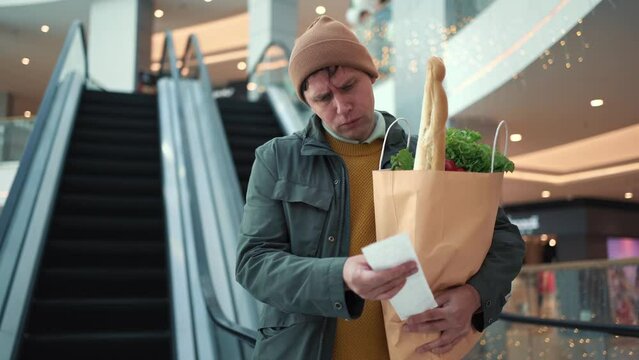 Price rise concept. Surprised male customer holding paper bag with food products looks at paper receipt after shopping in supermarket.