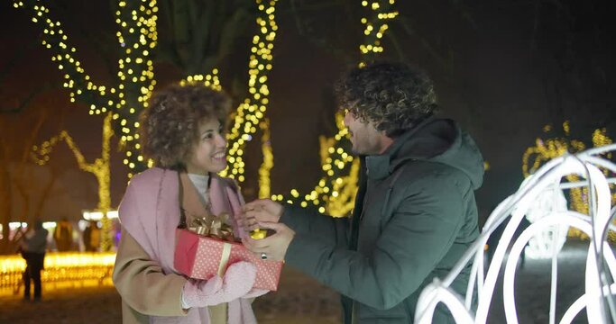Young African American woman makes a gift to a Indian man on background of lights and garlands, New Year and Christmas concept