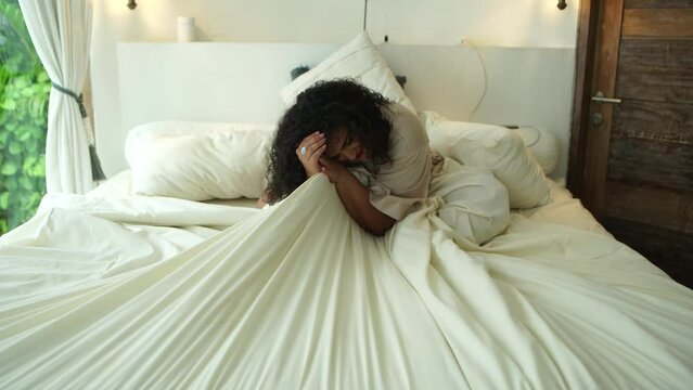 A middle-aged woman clutches the sheet tightly with her hands while sitting on the bed in the bedroom at home. The concept of trouble and despair in your personal life 4K