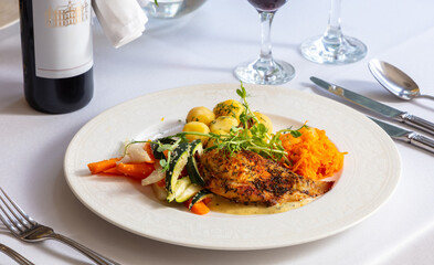 Herb-crusted chicken breast with boiled potatoes and glazed vegetables served on an elegant plate with a glass of red wine and sparkling water