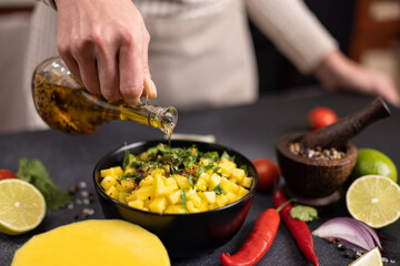 Cooking mango salsa - woman chef pouring cooking olive oil onto sliced chopped ingredients in a black ceramic bowl