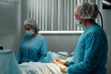 Obraz premium Experienced clinician in protective mask, gloves and medical scrubs using probe during surgical operation on leg or knee of patient