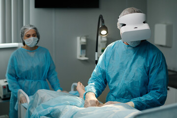 Experienced surgeon in VR headset performing surgical operation on leg of patient covered with...